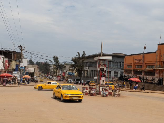 Ein gelbes Taxi fährt eine belebte Stadtstraße entlang, die von Gebäuden gesäumt ist, während Fußgänger mit Regenschirmen auf dem Gehweg unter wolkenverhangenem Himmel gehen.