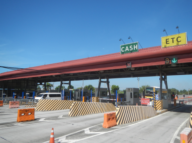 Eine Mautstation mit einem "Bar"-Schild, umgeben von Fahrzeugen, Barrieren, Schildern, Straßenlaternen, Strommasten, Stromkabeln, Bäumen und einem bewölkten Himmel.