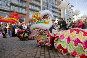 Ein lebendiges chinesisches Neujahrsfest in Amsterdam mit einer Löwen-Tanz-Show vor einer Zuschauermenge, einige machen Fotos, vor dem Hintergrund von Gebäuden, Laternenmasten und einem klaren blauen Himmel.
