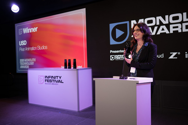 Frau in schwarzem Kleid, die auf einem Podium steht und einen Preis hält, mit einem Mikrofon und anderen Gegenständen auf dem Podium, einem Werbeplakat und einer Lampe im Hintergrund.
