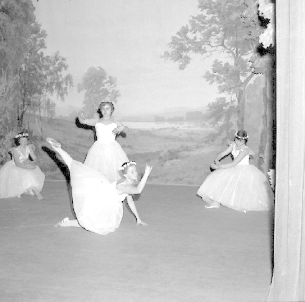 Vier junge Balletttänzerinnen in weißen Kleidern tanzen auf einer Bühne mit einer gemalten Kulisse aus Bäumen und Himmel in Schwarzweiß.