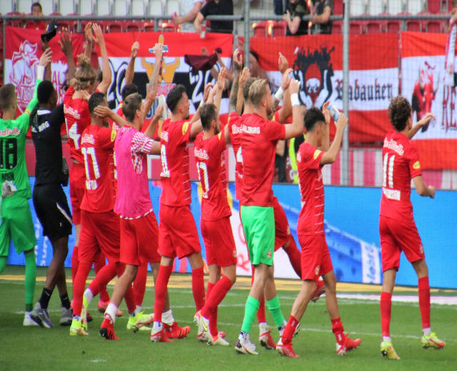Eine Gruppe von Fußballspielern feiert ein Tor auf einem Feld, einige halten Flaschen oder klatschen, mit Bannern, einem Zaun, Metallstangen, Stühlen und einem Dach mit Lampen im Hintergrund.