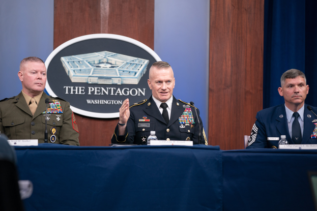 Drei Militärangehörige in Uniform sitzen an einem Tisch mit Namensschildern, Flaschen und Mikrofonen und diskutieren mit einem Verteidigungsminister im Hintergrund, der ein Logo und einen Vorhang zeigt.