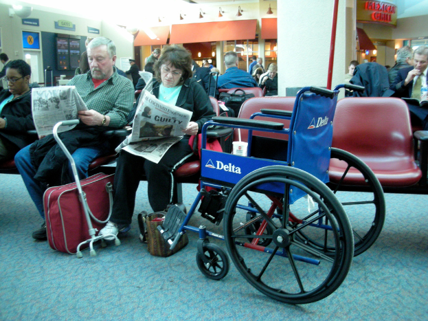 Eine Gruppe von Menschen, die auf Stühlen sitzen, wobei zwei Personen Zeitungen halten, ein Rollstuhl im Vordergrund und ein Balken mit Lichtern in der Mitte.