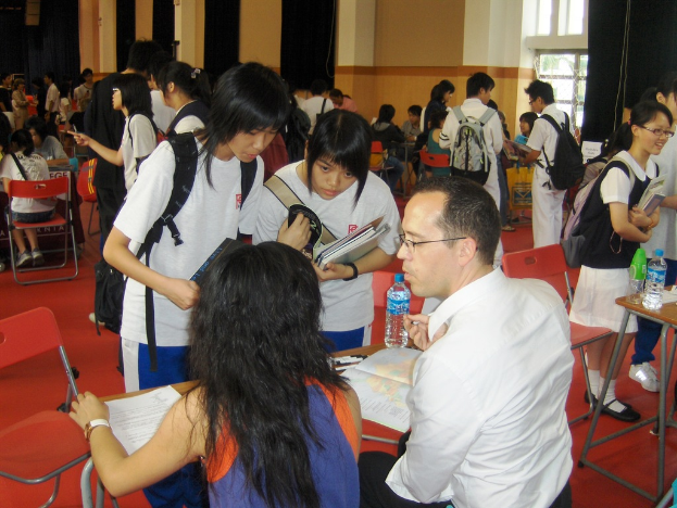 Ein Klassenzimmer mit mehreren Mädchen in weißen T-Shirts, die Bücher in der Hand halten, vorne, ein Mann und eine Frau im Vordergrund, die diskutieren, und eine gelbe Wand mit einem Glasfenster im Hintergrund.