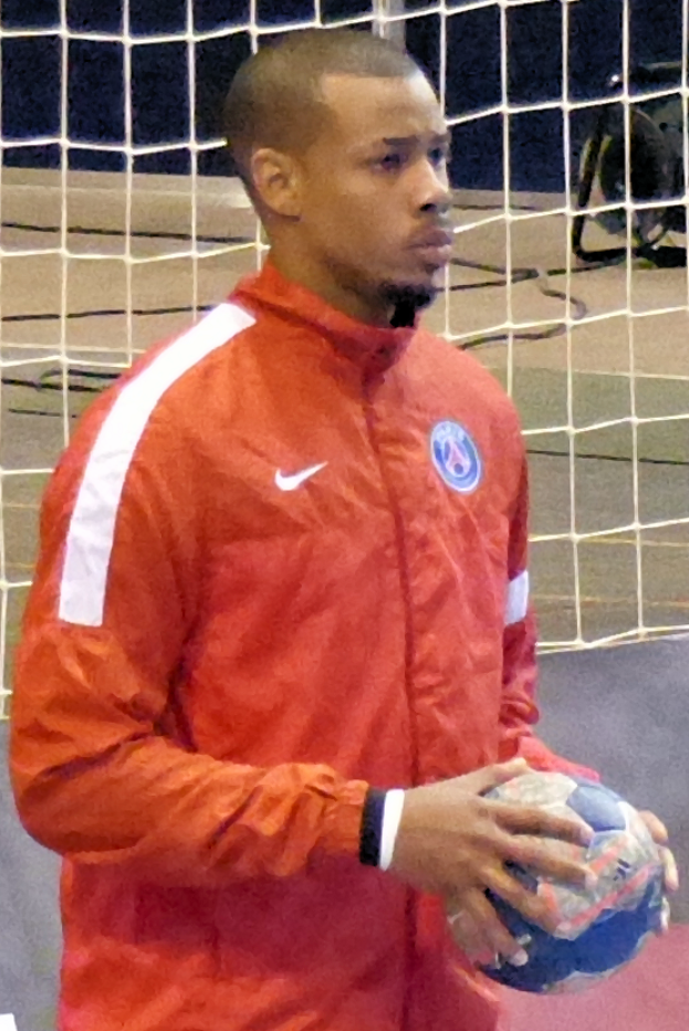 Ein Mann in einem blauen Hemd und Shorts steht vor einem Tor und h├Ąlt einen Fussball mit entschlossener Miene.