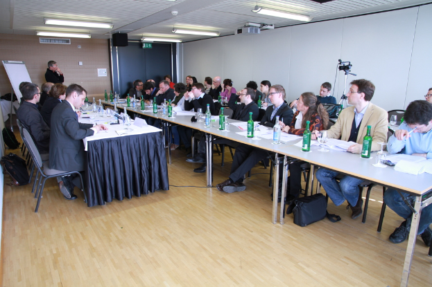 Ein Büroraum mit Mitarbeitern an Tischen sitzend, Rucksäcke auf dem Boden, ein Mann im Hintergrund und verschiedene Gegenstände wie Flaschen, Papiere, eine Kamera, Lautsprecher, Schilder, Projektor und Lampen.