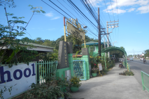 Der Eingang einer Schule mit einem Schild, das "Schule" auf der Seite einer Straße trägt, mit einem Bogen, Tor, Namensschild, Pflanzen, Bäumen, Strommasten mit Drähten und einem bewölkten Himmel im Hintergrund.