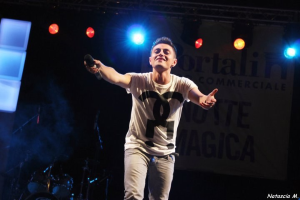 Ein Mann auf einer hell erleuchteten Bühne hält ein Mikrofon, mit Musikinstrumenten und einem "Nouveau Magica"-Schild im Hintergrund.