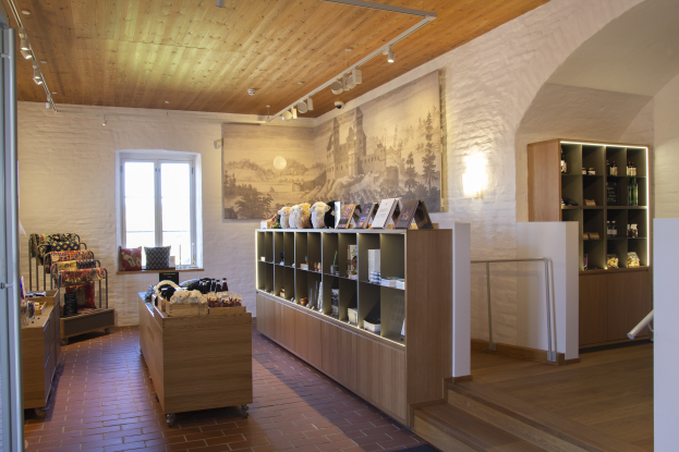 Ein unordentlicher Raum in einem Laden in Heidelberg, Deutschland, mit Möbeln, Regalen mit Gegenständen, einem gerahmten Foto an der Wand, einem Fenster, Deckenleuchten und einer Treppe.