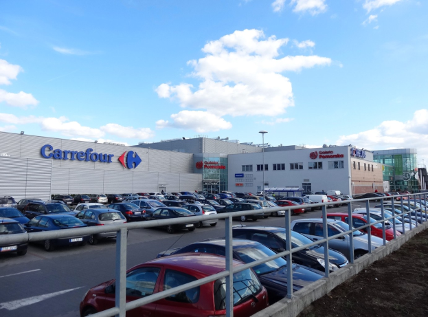 Großes Autohaus mit zahlreichen geparkten Fahrzeugen, umgeben von städtischer Infrastruktur wie Straßenlaternen, Gebäuden und einem bewölkten Himmel.