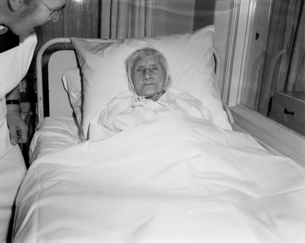 Alte Frau im Krankenhausbett mit einer Krankenschwester daneben, Bett mit einem weißen Laken und Kissen bedeckt, Fenster mit Vorhängen im Hintergrund.