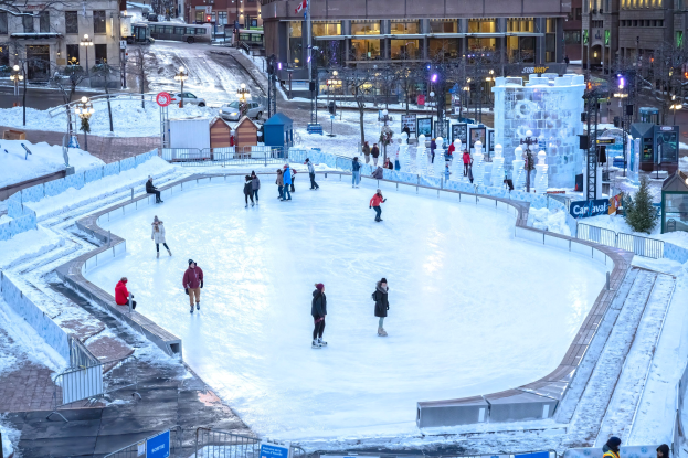 Eine Gruppe von Menschen, die auf einer Eisbahn in einer Stadt skaten, umgeben von Geländern, Schildern, Laternen, Bäumen und Gebäuden mit Fenstern, mit Fahrzeugen auf der Straße und einer Fahne mit einem Mast im Hintergrund.
