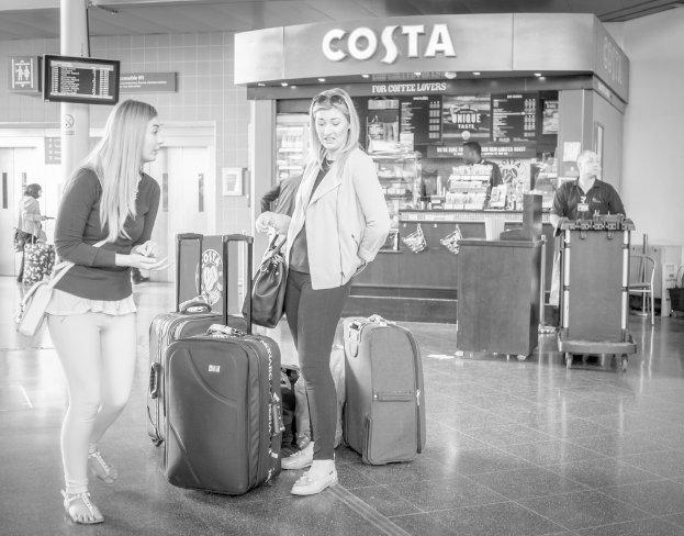 Zwei Frauen vor mehreren Gepäckstücken mit einem Kaffeehaus im Hintergrund, wahrscheinlich am Flughafen.
