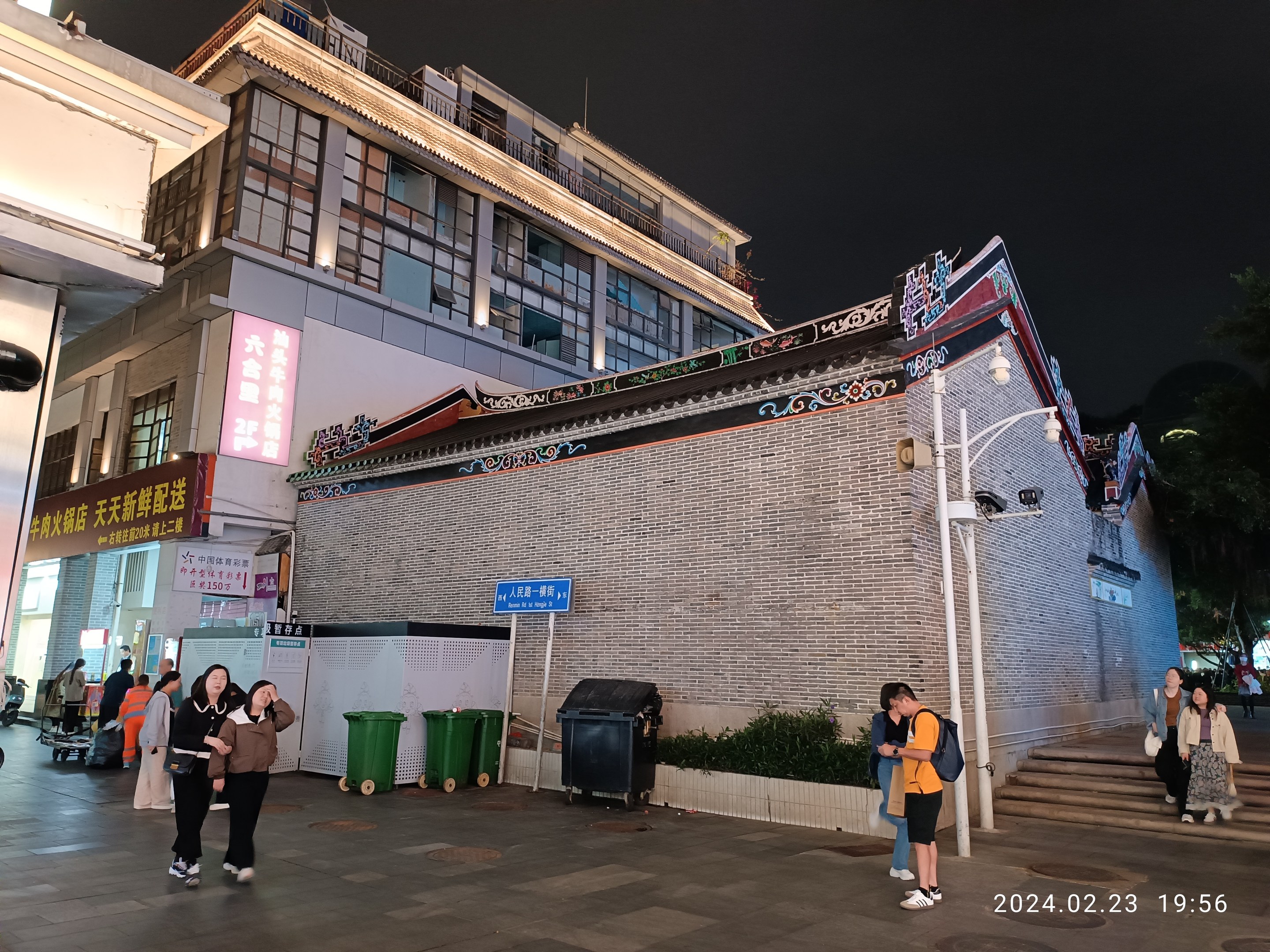 Menschen gehen nachts eine Straße entlang mit Gebäuden, Straßenlaternen, Schildern, Pflanzen und Bäumen.