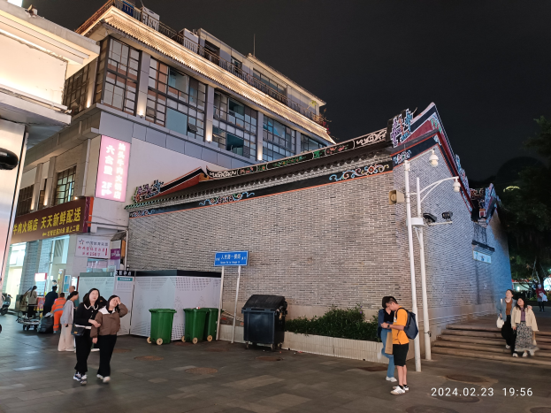 Menschen gehen nachts eine Straße entlang mit Gebäuden, Straßenlaternen, Schildern, Pflanzen und Bäumen.