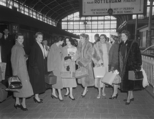 Schwarz-weißes Bild von Frauen, die an einem Bahnhof stehen, links ein Zug und rechts Geländer und eine Tafel, jede hält eine Tasche.