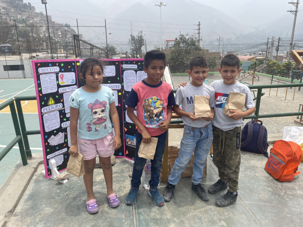 Gruppe von Kindern auf einem Basketballfeld mit Papiertüten in den Händen, Tüten und Gegenstände rechts auf dem Boden und eine Tafel mit Text links, vor einem Hintergrund aus Zaun, Pfosten, Straßenlaternen, Bäumen, Gebäuden, Hügeln und Himmel.