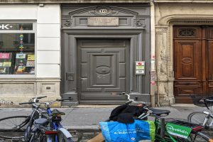 Mehrere Fahrräder sind in einer Reihe auf dem Gehweg vor einem Gebäude mit einer Tür und einem Fenster auf der linken Seite geparkt.