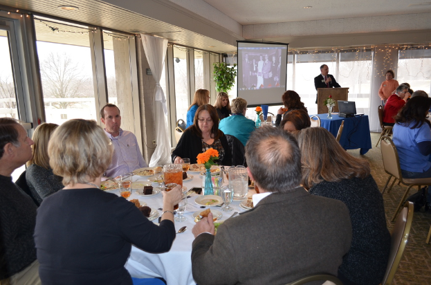 Eine Gruppe von Menschen sitzt um einen Tisch mit verschiedenen Speisen, während eine Person steht und spricht, ein Projektor zeigt ein Bild, eine Pflanze daneben und ein Blick aus dem Fenster, Himmel und Bäume im Hintergrund.