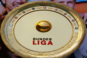 Goldener Schild mit "Bundesliga"-Text in der Hand eines Mannes, Hintergrund zeigt eine Fahne mit drei Personenbildern.