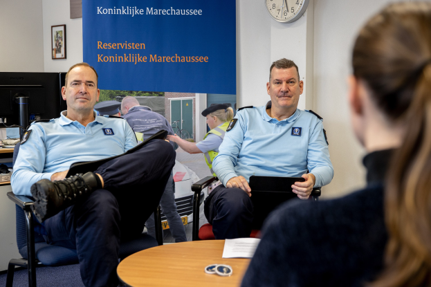 Zwei Polizeibeamte sitzen an einem Tisch mit einer Frau, die Dokumente durchsieht, mit einer Wanduhr und einem Banner im Hintergrund, einem Computer-Setup auf einem benachbarten Tisch und gerahmten Fotos an der Wand.