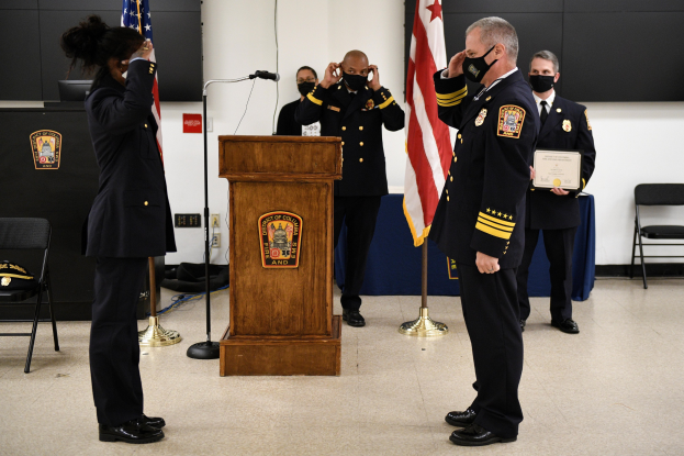 Gruppe uniformierter Personen in Masken vor einem Rednerpult während einer Vereidigung eines neuen Feuerwehrchefs, der eine Urkunde hält, mit Flaggen, Stühlen, Tischen und einer Wand im Hintergrund.