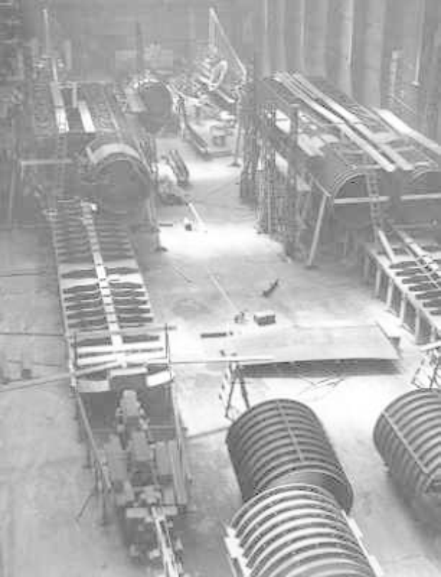 Schwarzes und weiße Foto eines industriellen Fabrikinnern mit zahlreichen Rohren, Maschinen und Ausrütung auf dem Boden, das stützende Pfeiler und eine ferne Wand zeigt.