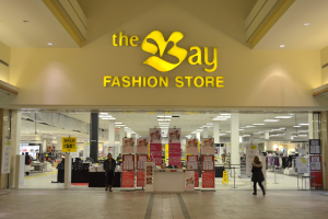 Außenansicht eines Bay Fashion Stores in einem Einkaufszentrum mit Einkäufern, Wandtexttafeln, Säulen, Deckenleuchten und Hintergrundauslagen.
