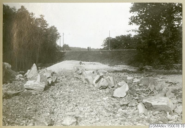 Schwarz-weiß-Foto einer Schotterstraße mit verstreuten Steinen, Bäumen, Strommasten, einer Wand im Hintergrund und dem Himmel darüber, mit Text, der Routen am unteren Rand auflistet.