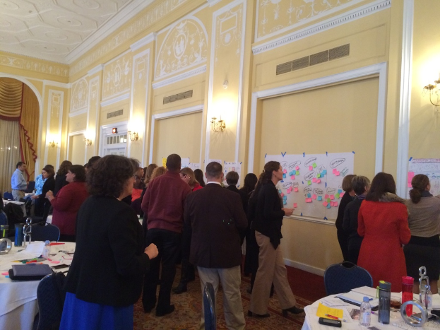 Eine Gruppe von Menschen betrachtet Plakate und Charts, die an einer Wand in einem Raum zu sein scheinen, der wie ein Kunstmuseum aussieht.