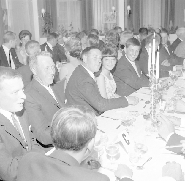 Eine Gruppe von Menschen sitzt an einem langen Tisch, lächelt und genießt die Gesellschaft des anderen, mit Gläsern, Besteck, Kerzen und anderen Gegenständen auf dem Tisch, vor einem Hintergrund aus Vorhängen und Wandlampen.
