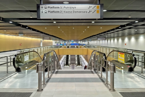 Ein leerer U-Bahnhof mit Rolltreppen, Geländern, Texttafeln, Deckenleuchten und Wänden, mit ein paar Menschen, die reges Treiben vermuten lassen.