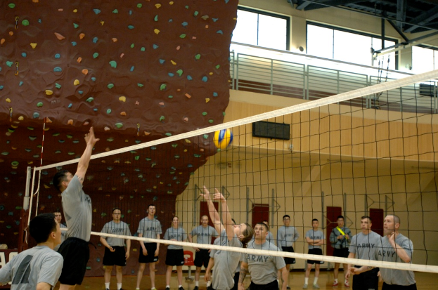 Eine Gruppe von Menschen in Kleidung, Socken und Schuhen spielt Volleyball auf einem Court mit Netz, Antennen, einem Zaun und Fenstern im Hintergrund.