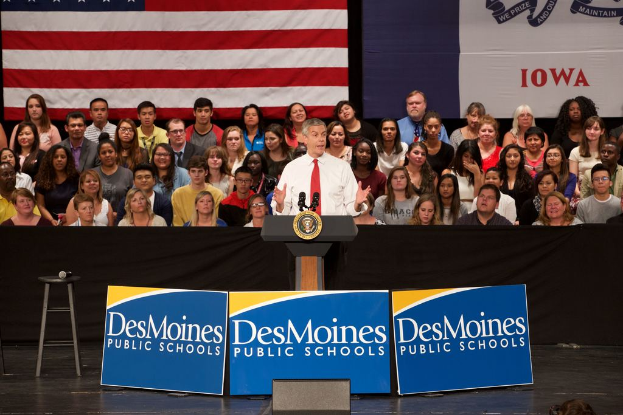 Ein Mann in einem weißen Hemd und roter Krawatte steht an einem Podium mit drei Tafeln, auf denen Text steht, einem Hocker mit Mikrofon und einem sitzenden Publikum im Hintergrund, mit einer Flagge und zusätzlichem Text.