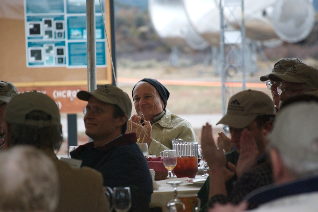 Menschen, die auf Stühlen sitzen und klatschen, mit einem Tisch daneben, der Gläser, einen Teller und ein Tuch enthält, sowie einem Pfahl, einer Tafel und Gras im Hintergrund.