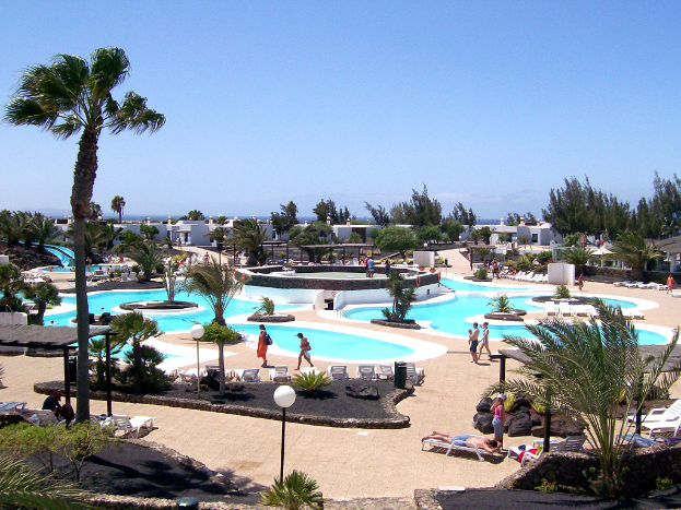 Ein Resort mit Pools, umgeben von Bäumen, Menschen zwischen den Pools spazieren, Gebäude im Hintergrund und ein klarer Himmel darüber.