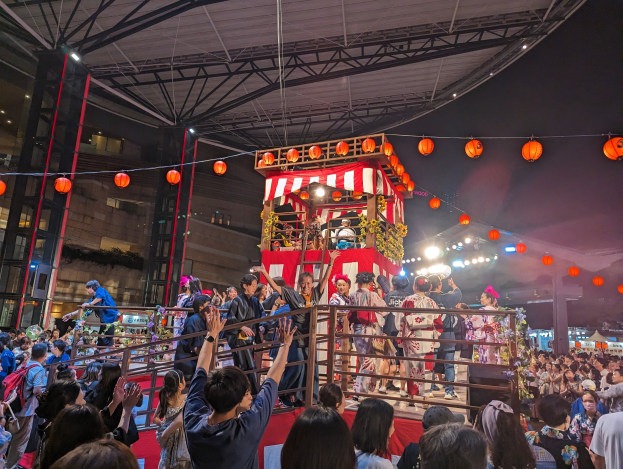Eine Menschenmenge steht auf einer beleuchteten Bühne mit Laternen, einige halten Kameras, umgeben von einem Geländer, mit Bäumen und einem klaren blauen Himmel im Hintergrund bei Nacht.