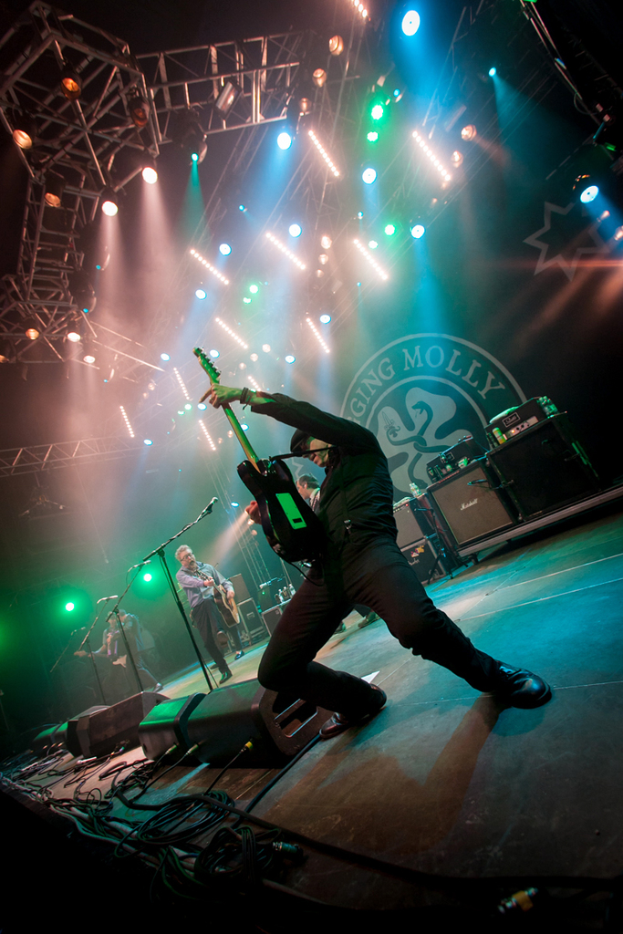 Ein musikalisches Konzert mit drei Performern auf der Bühne: zwei Gitarristen rechts mit Mikrofonen davor, ein Hintergrund mit dem Wort "MOLLEY" und zwei Schlangensymbolen sowie Scheinwerfer darüber.