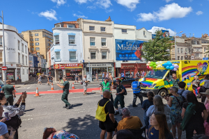 Eine Gruppe von Menschen steht vor einem Rettungswagen auf einer Straße, umgeben von Absperrbaken, Bäumen, Gebäuden mit Fenstern, Laternen und einem Banner mit Text, unter einem bewölkten Himmel bei der London Pride Parade.