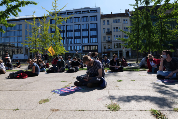 Eine Gruppe von Menschen sitzt vor einem Gebäude auf dem Boden während einer Demonstration in Berlin, einige tragen Masken und Taschen sind verstreut, vor Bäumen und einem klaren blauen Himmel.