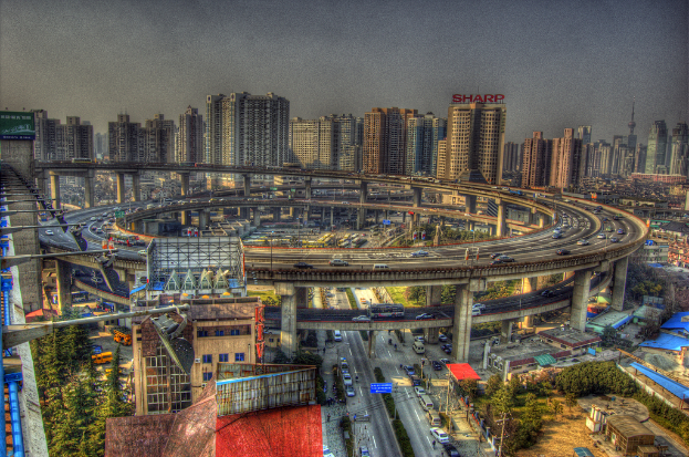 Stadtpanorama mit einer zentralen Ringstraße mit Fahrzeugen, einer unteren Straße mit Autos und Fahrrädern, zahlreichen Hochhäusern im Hintergrund, einem Himmel oben, Bäumen auf der linken Seite unten und kleinen Häusern, die über die Stadt verstreut sind.