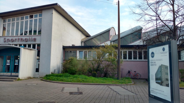 Ein großes Gebäude mit einem Schild davor, umgeben von Bäumen, Pflanzen, Gras, Fahrrädern und einem Pfahl, mit einem bewölkten Himmel im Hintergrund, als Sportballe identifiziert, zur Eröffnung 2020 vorgesehen.