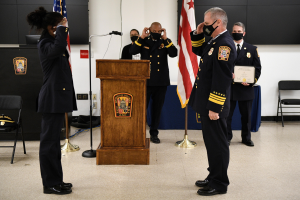 Gruppe uniformierter Personen in Masken vor einem Rednerpult bei einer Vereidigung eines neuen Feuerwehrchefs, der eine Urkunde hält, mit Fahnen, Stühlen, Tischen und einer Wand im Hintergrund.
