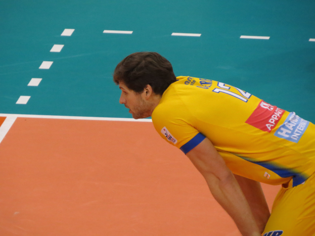 Ein professioneller Volleyballspieler in einem gelben T-Shirt steht selbstbewusst auf einem Court und bereitet sich auf den Aufschlag vor.