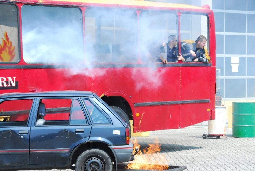 Roter Doppeldeckerbus mit Rauch aus dem Auspuff, daneben geparktes Auto, drei sichtbare Insassen und ein Gebäude mit Glasfenstern und einem Fass im Hintergrund.