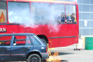 Roter Doppeldeckerbus mit Rauch aus dem Auspuff, daneben geparktes Auto, drei sichtbare Insassen und ein Gebäude mit Glasfenstern und einem Fass im Hintergrund.
