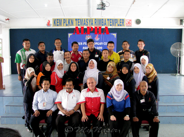 Eine Menschenmenge, die fröhlich posiert und vor einer Bühne fotografiert wird, mit einer Wandtafel dahinter, einem Tischventilator rechts, einem Podium links und einem Projektor oben im Bild.