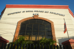 Das Gebäude des Departments für Soziales und Entwicklung mit Text an der Wand, einem Fahnenmast mit wehender Flagge, einem Metallzaun, Pflanzen, Bäumen, Drähten und einem bewölkten Himmel.
