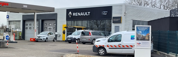 Ein Renault-Autohaus mit parkenden Autos davor, umgeben von einem Metallzaun, einem Schild, Pfählen, einer Baumgruppe und einem bewölkten Himmel.